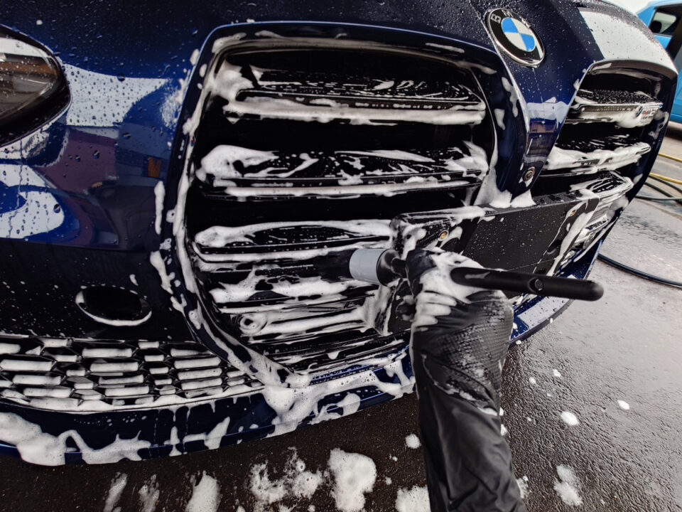 Blue BMW being washed with soap suds; a gloved hand holds a foam-wand near the front grille.