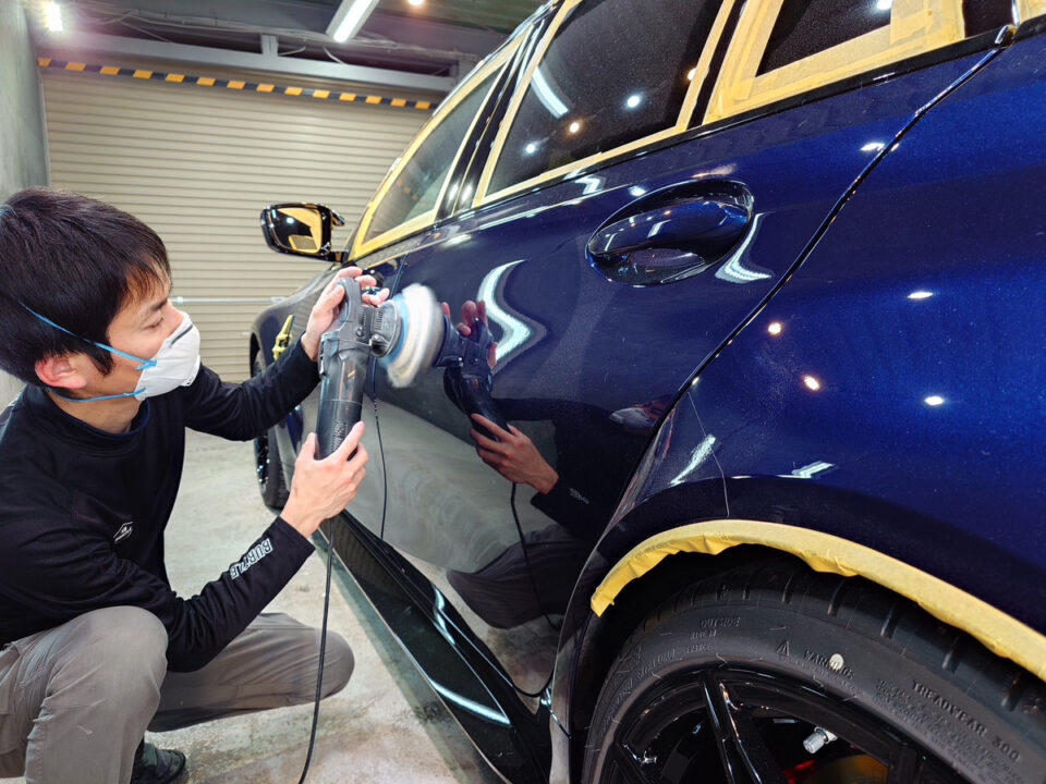 Technician wearing a mask polishes a dark blue car with a power buffer in a workshop, protective tape along windows and edges.
