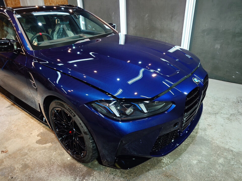 Blue BMW sedan in an indoor showroom, front-left three-quarter view with glossy finish and reflections from overhead lights.