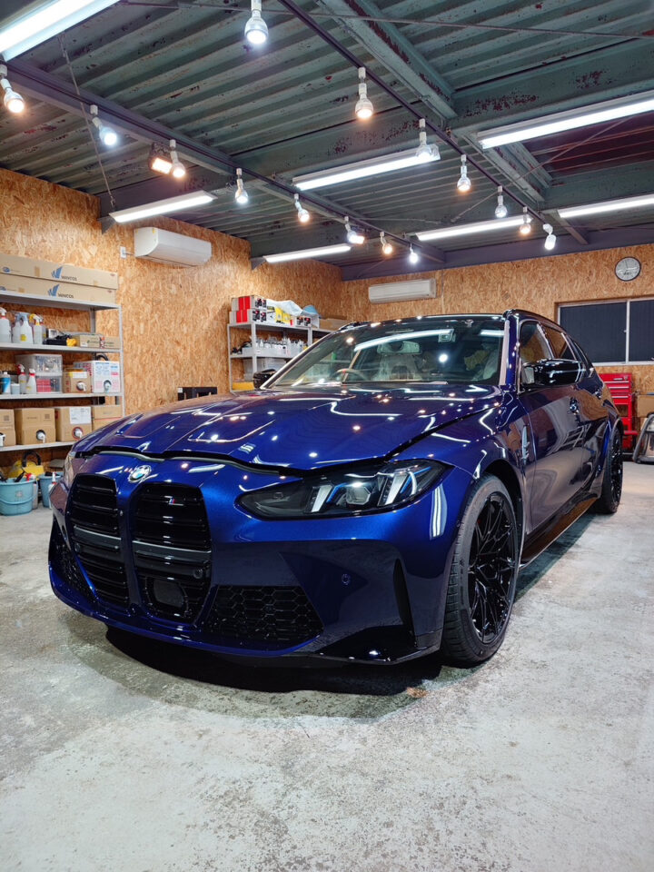 Blue BMW M car parked inside a workshop, with shelves of supplies and a concrete floor visible around it.
