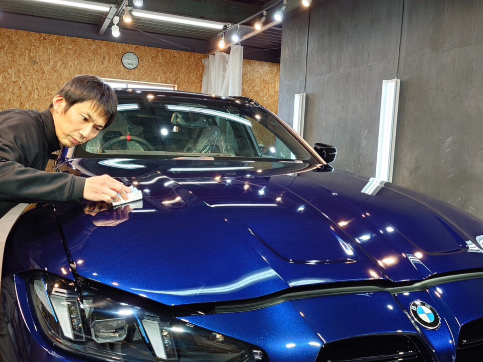 Man polishing a metallic blue BMW in a workshop, using a cloth on the car's hood under bright studio lights