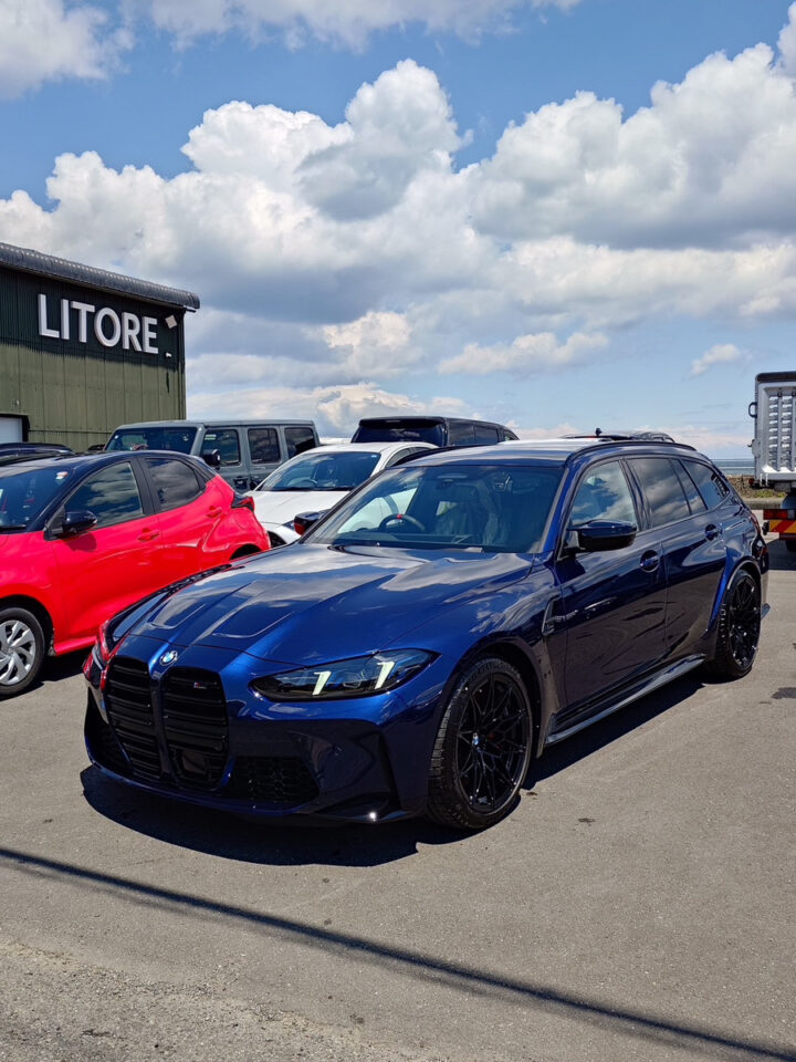 Blue BMW sedan with black wheels in a parking lot beside a red car under a cloudy sky.