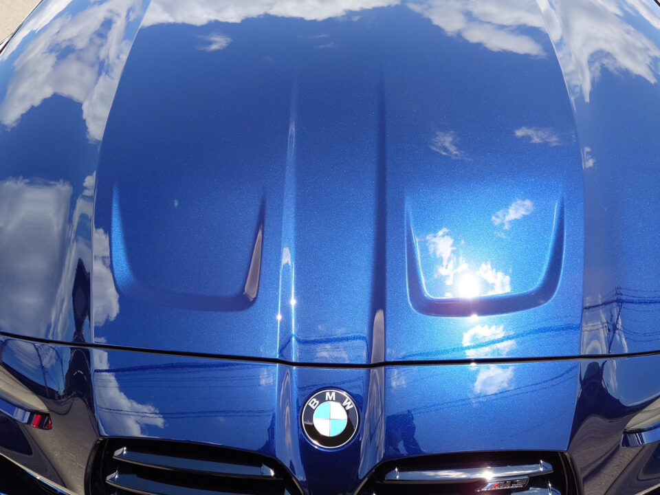 Blue BMW hood with emblem, reflections of clouds and sky on glossy surface.