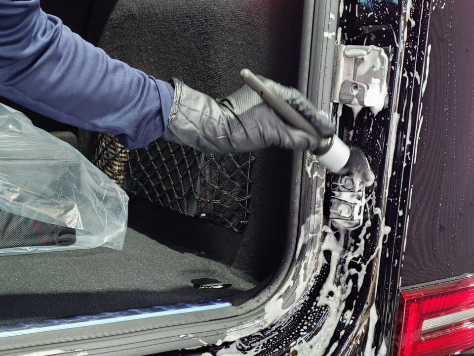 Person wearing gloves scrubs the edge of a car trunk with soapy foam and a sponge brush inside the open cargo area.