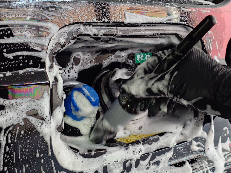 Person wearing a glove scrubs the car's engine bay with soapy foam and a brush.