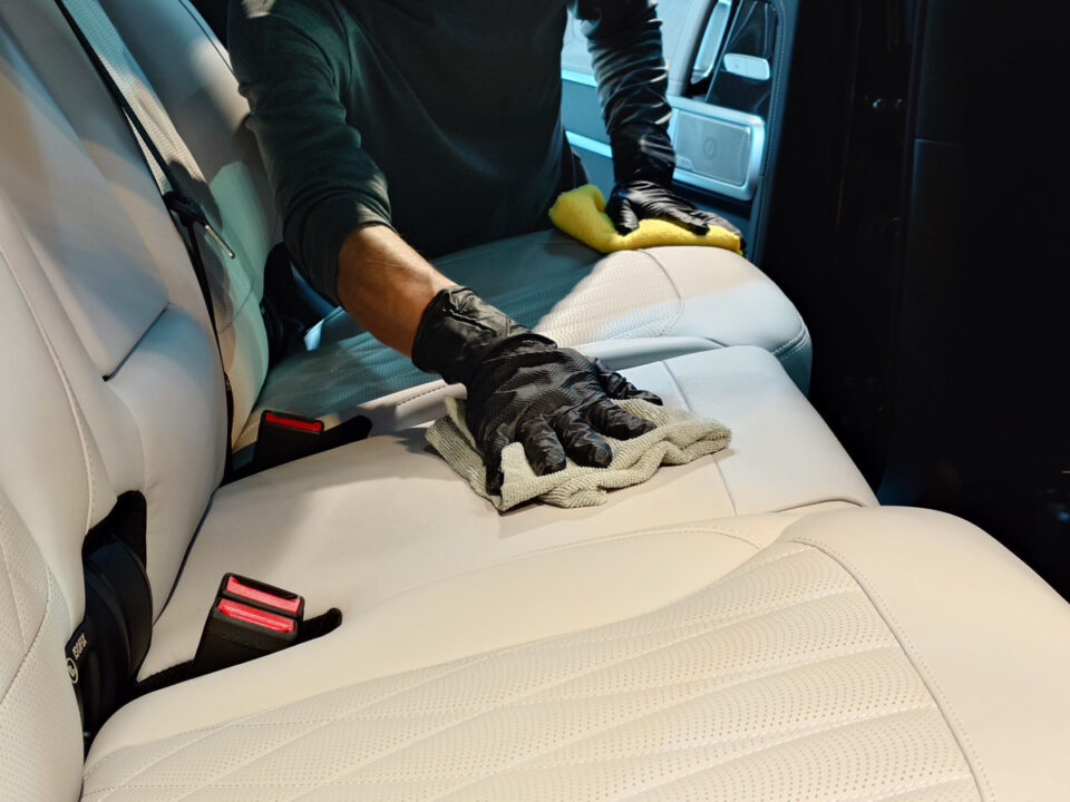 Person wearing black gloves scrubbing a white leather car seat with a yellow sponge in the car interior.