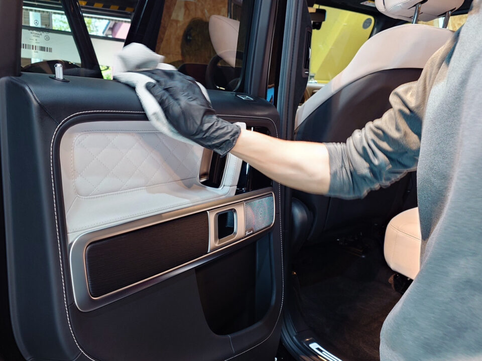 Person wearing a black glove cleans the interior door panel of a car with a cloth, showing quilted trim and controls on the door.
