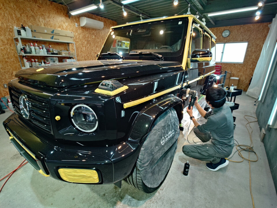 Technician kneels beside a black Mercedes G-Class, polishing the front fender with a power tool in a well-lit workshop.