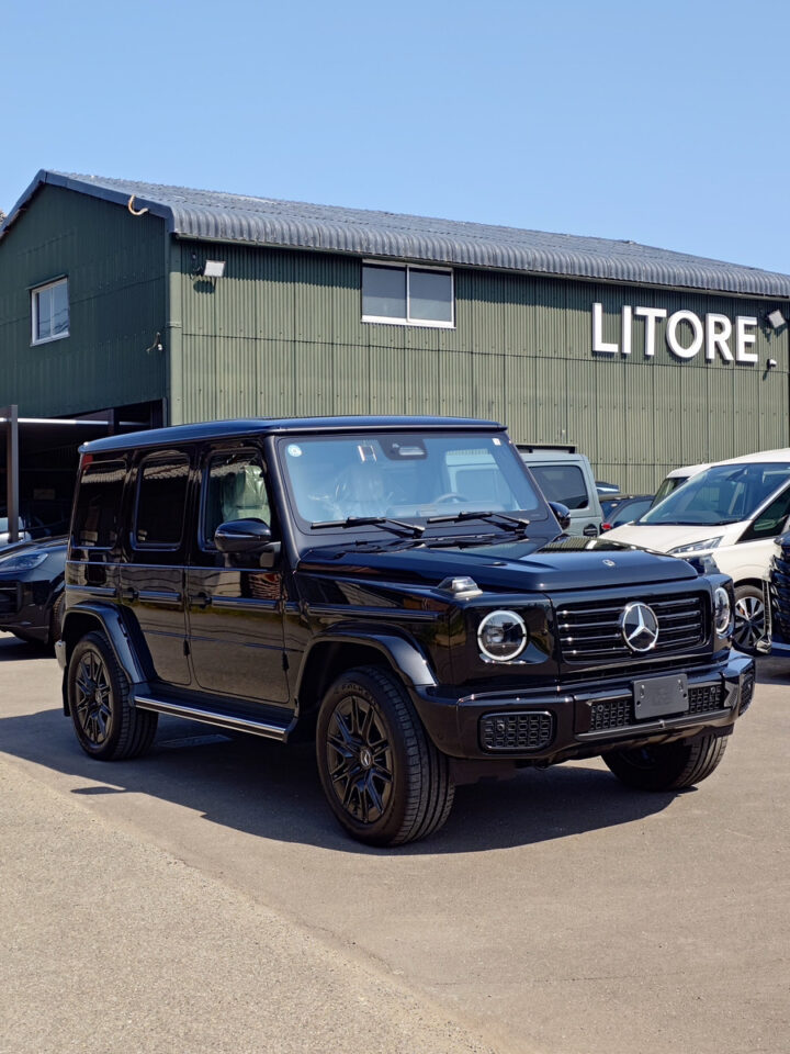 Black Mercedes-Benz G-Class SUV parked in a dealership lot in front of a green industrial building with a large 'LITORE' sign and other vehicles nearby.
