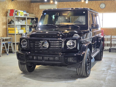 Front view of a black Mercedes-Benz G-Class SUV parked inside a workshop with shelves of supplies in the background.