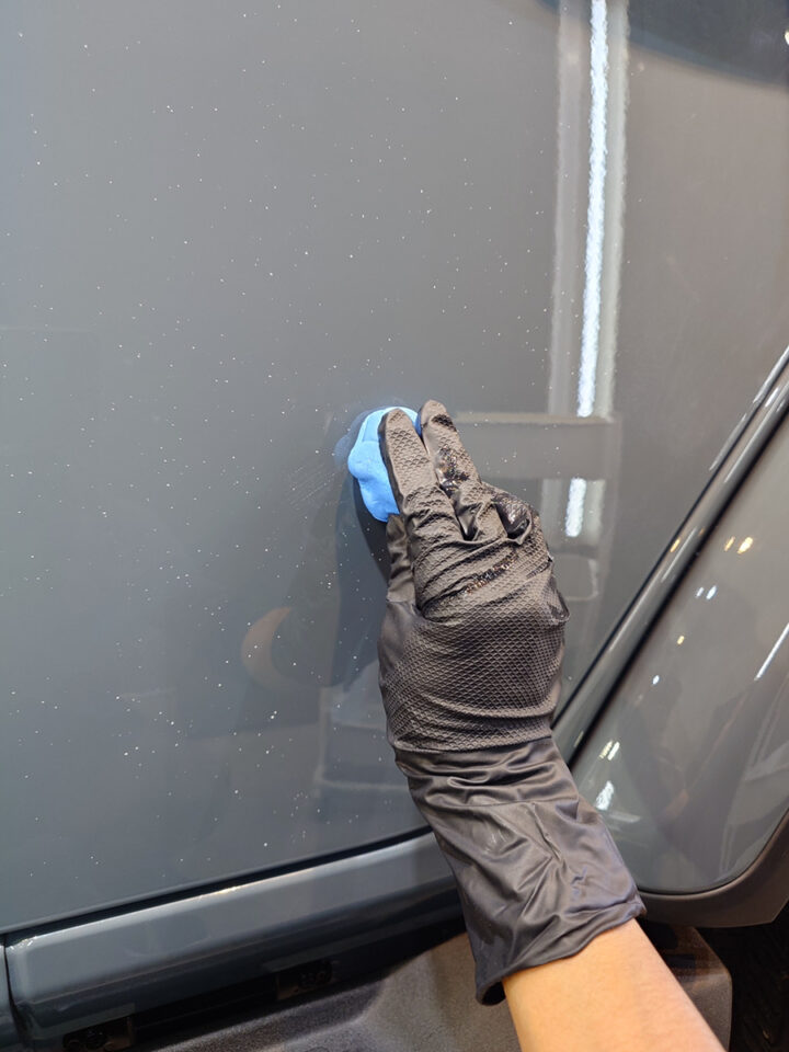 Gloved hand buffing a gray car door with a blue microfiber cloth during detailing edge of window visible