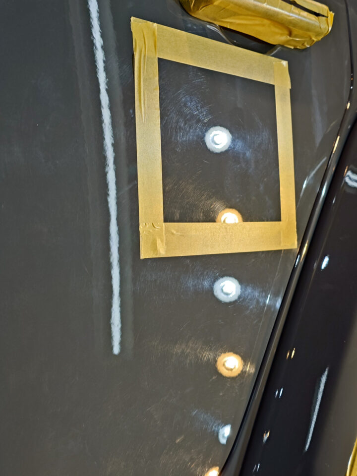 Close-up of a dark car door with masking tape forming a square around a repair area and ceiling light reflections on the glossy surface.