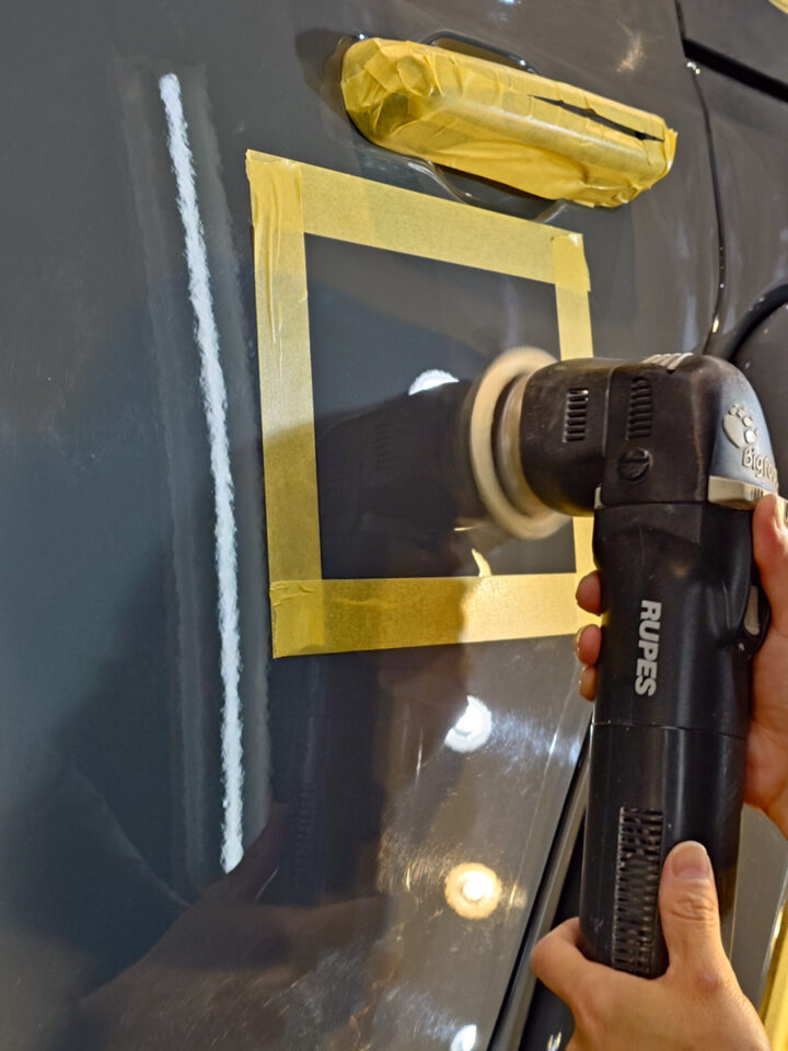 Person using a polishing tool on a car door, masking tape square around a panel for paint work.