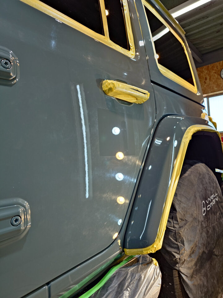 Close-up of a gray SUV door being repainted, masking tape around windows and wheel arch, tire protected with a cover.