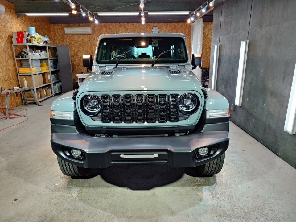 Front view of a light blue Jeep Wrangler inside a workshop, with shelves and tools in the background.