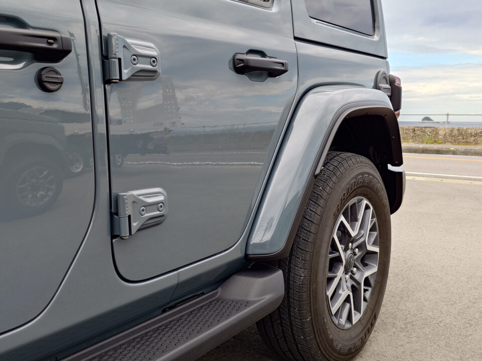 Close-up of a gray Jeep Wrangler's front door, hinge, and wheel with fender flare and running board visible by the roadside