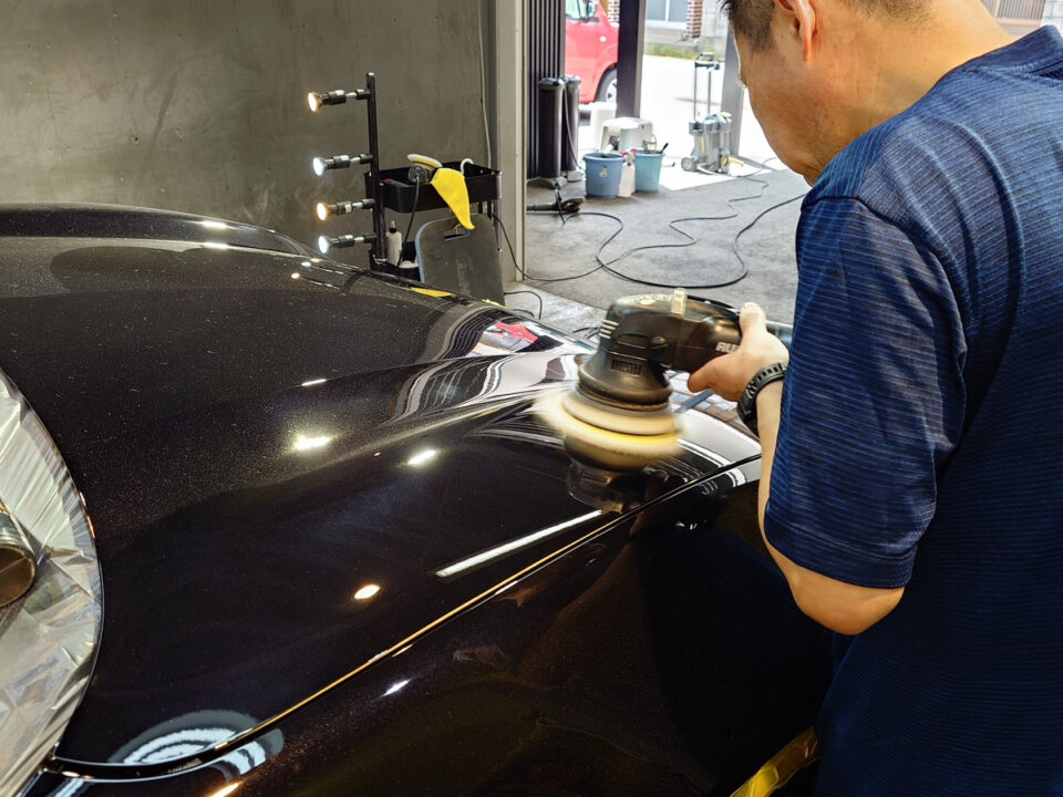 Person in a blue shirt buffing a dark car hood with a power polisher in a workshop.