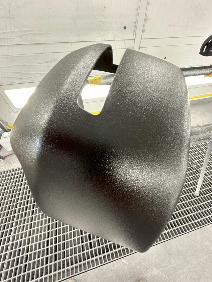 Large black textured plastic car interior panel with a central U-shaped opening, in a paint booth.