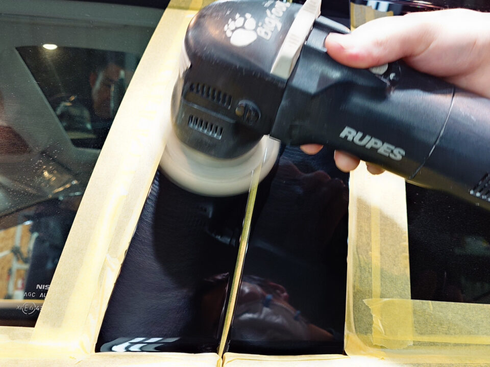 Close-up of a hand-held Rupes polisher buffing a car window with masking tape around the edges for protection.