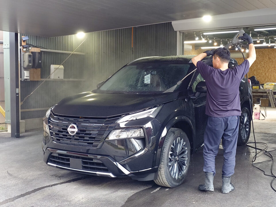 Worker washing a black Nissan SUV with a pressure washer in an indoor car wash bay.