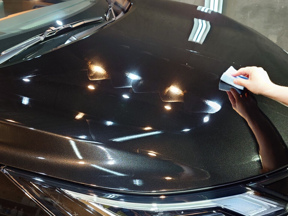 Person wiping a dark car hood with a blue cloth, polishing the surface under showroom lights.