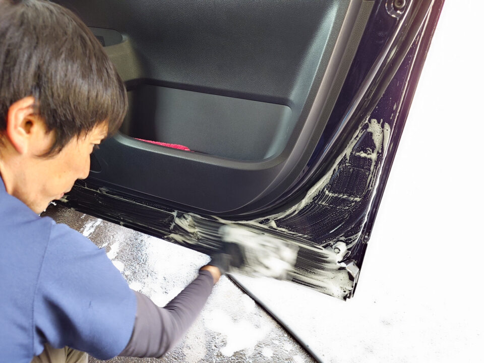 Person washing the bottom edge of a car door with soap and a sponge on a snowy surface.
