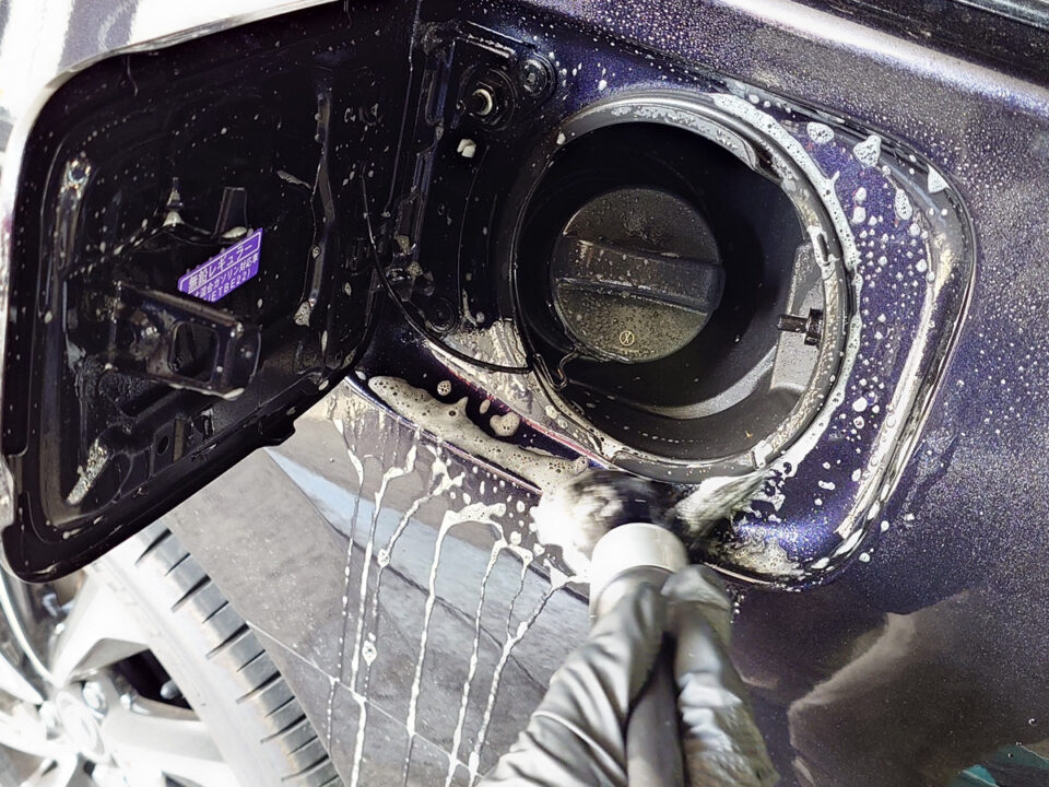 Close-up of a car's open fuel filler door with the gas cap exposed, while someone washes the area with soapy water.