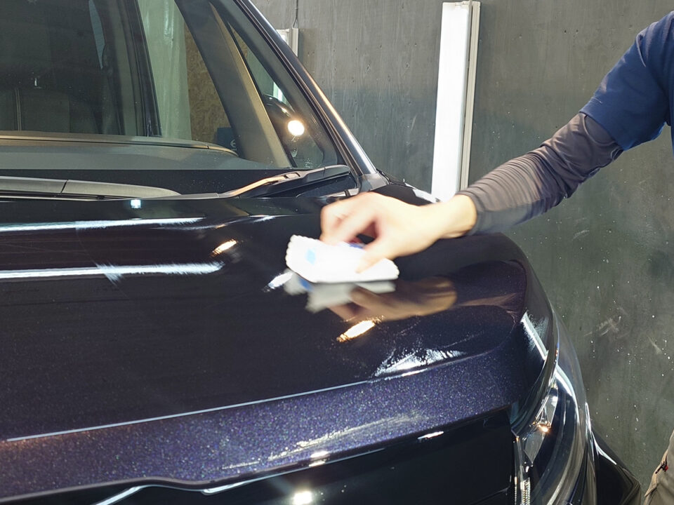 Person wiping the shiny dark-blue car hood with a cloth in a workshop, close-up on the hood and windshield area.