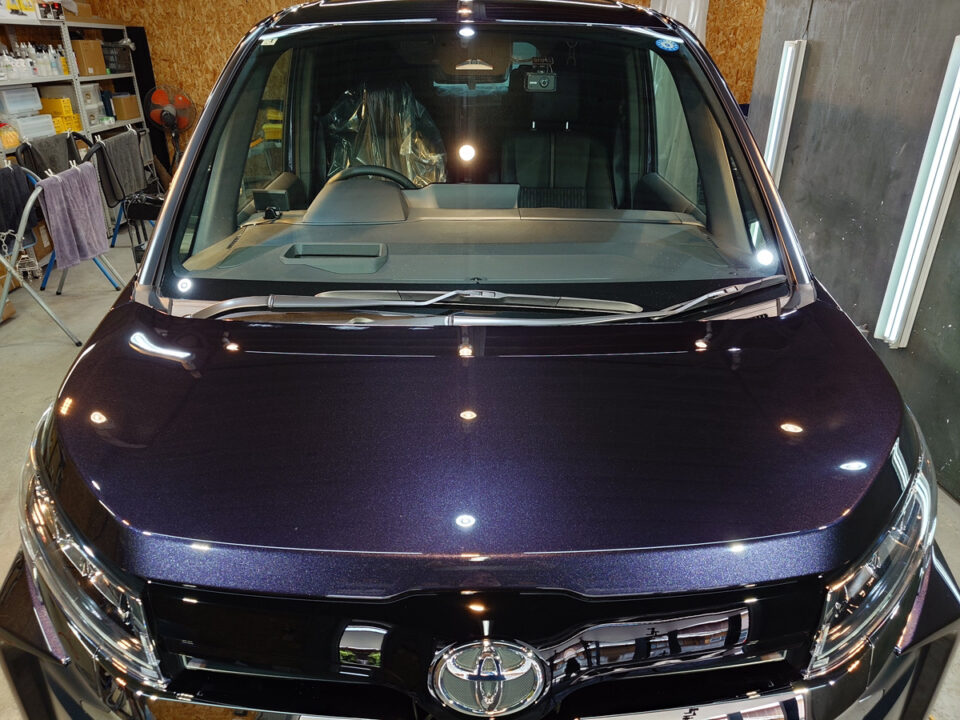 Front view of a dark purple Toyota SUV in a workshop, hood reflecting bright overhead lights.