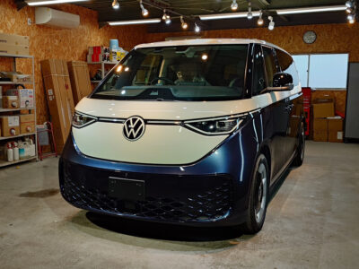Front view of a white-and-navy Volkswagen van parked inside a cluttered workshop with shelves, boxes, and tools in the background.