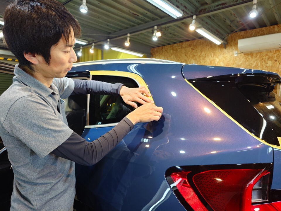 Person applying protective film to the rear side of a blue car in a workshop.