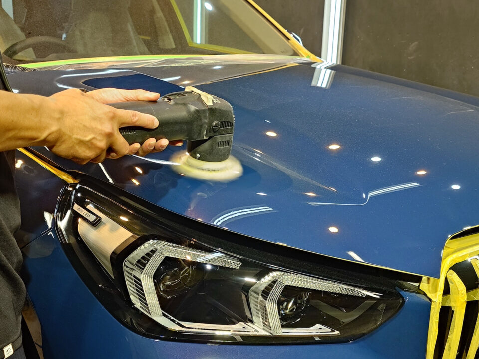 Person polishing a blue car hood with a power buffer during detailing, masking tape visible along the edges.