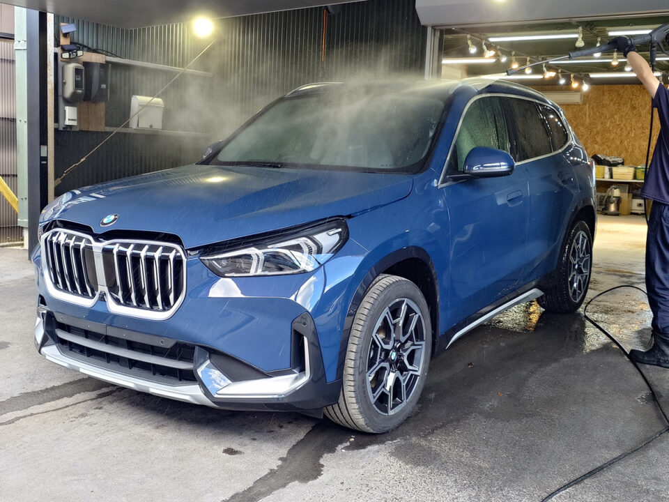 Blue BMW SUV being washed in a car wash bay, water spraying over the windshield as a technician uses a hose.