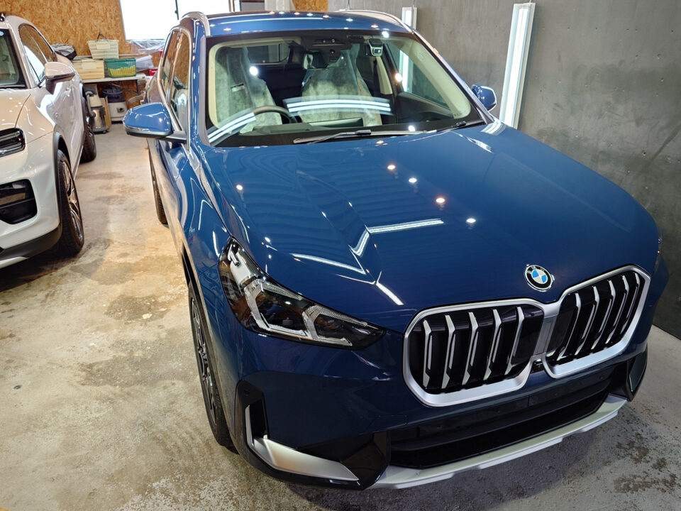 Front view of a blue BMW SUV parked in a garage with a white SUV nearby and workshop items in the background.
