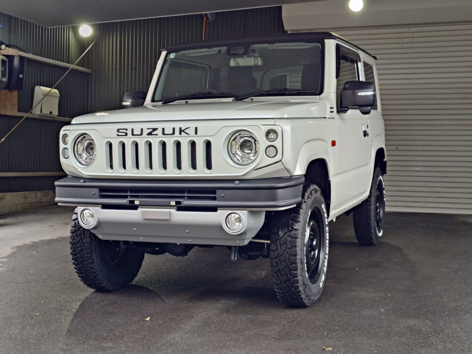 White Suzuki off-road SUV (Jimny) parked inside a garage, angled front view showing round headlights, seven-slot grille, and rugged tires.