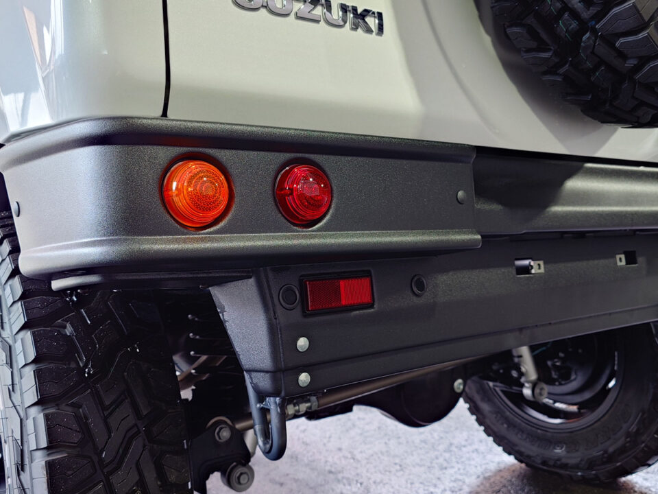 Rear of a Suzuki SUV showing two round taillights (orange and red) on a textured black bumper with a spare tire nearby.