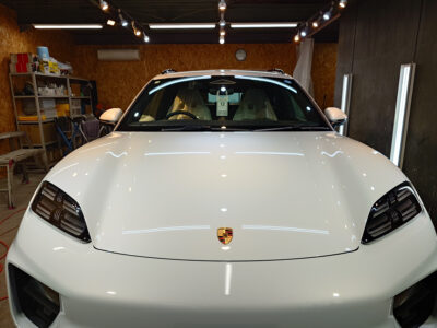 Front view of a white Porsche parked in a garage workshop with shelves and tools in the background.