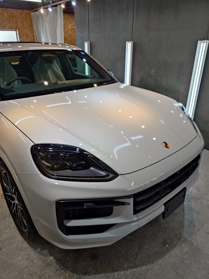 Front three-quarter view of a white Porsche SUV in a showroom, highlighting the sleek headlights and large front grille.