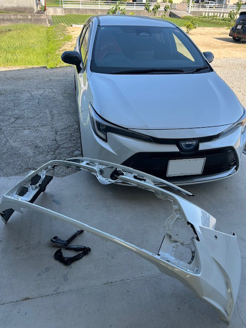 Front view of a white car with its front bumper removed, the bumper frame lying on the ground beside it in a parking area.